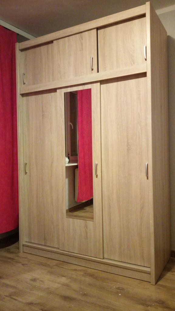 Jasna, drewnopodobna szafa przesuwna z lustrem w centralnej części, uzupełniona nadstawką z dodatkowymi szafkami, ustawiona na panelach podłogowych, z widocznym fragmentem czerwonej zasłony w tle.