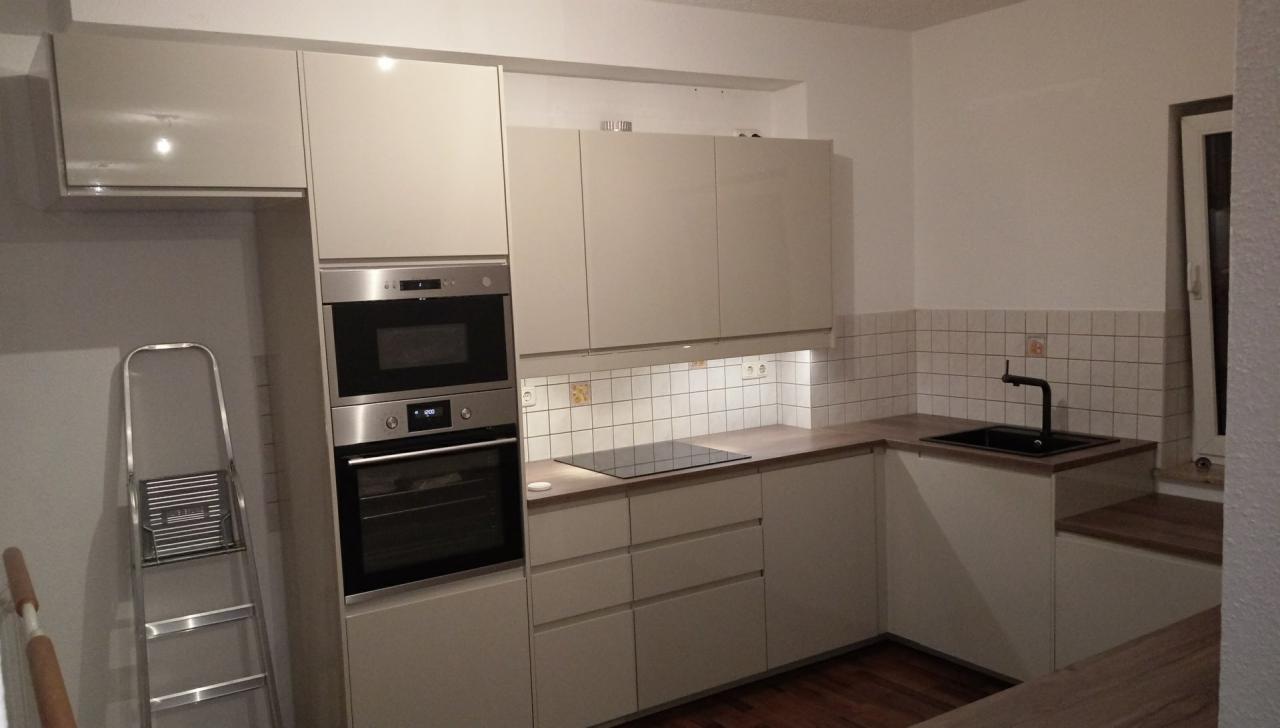 Jasna kuchnia z białymi szafkami, piekarnikiem, płytą indukcyjną i czarną armaturą, widoczna drabina aluminiowa w rogu.