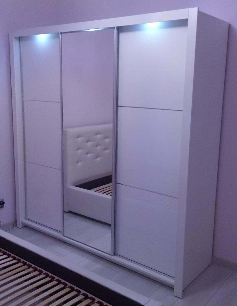 Biała szafa przesuwna z lustrem w sypialni, widoczna rama łóżka i zagłówek z pikowaniem, oświetlenie LED na górze szafy.