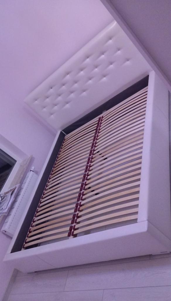 Białe łóżko z tapicerowanym zagłówkiem w trakcie montażu, widoczne drewniane listwy stelaża i fragment ściany z grzejnikiem.