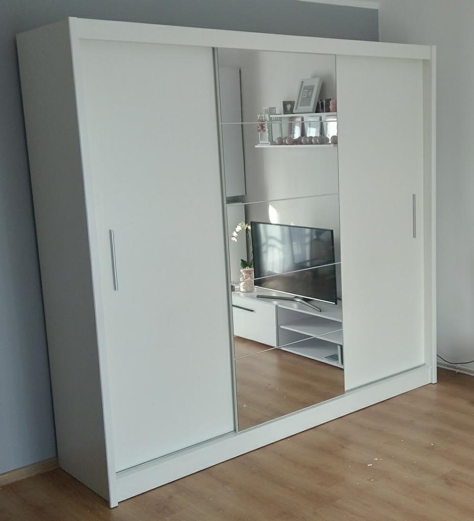 Biała szafa przesuwna z lustrzanymi drzwiami, odbijająca telewizor i półki, stojąca na drewnianej podłodze.