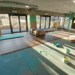 Kamil Kowalski - Wnętrze w trakcie prac, widoczne przygotowane panele podłogowe, puszki z farbą, płyty OSB i materiały izolacyjne, duże okna z widokiem na zewnątrz.