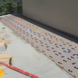 Układanie szarych płytek na tarasie z użyciem pomarańczowo-niebieskich klipsów poziomujących. Widoczna poziomica i kartony z płytkami.