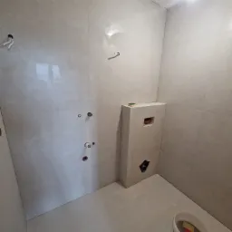 Jasna łazienka po remoncie z beżowymi płytkami, widoczne przyłącza wodne, fragment toalety podtynkowej i wiadro z gąbkami. Ukończone prace wykończeniowe.