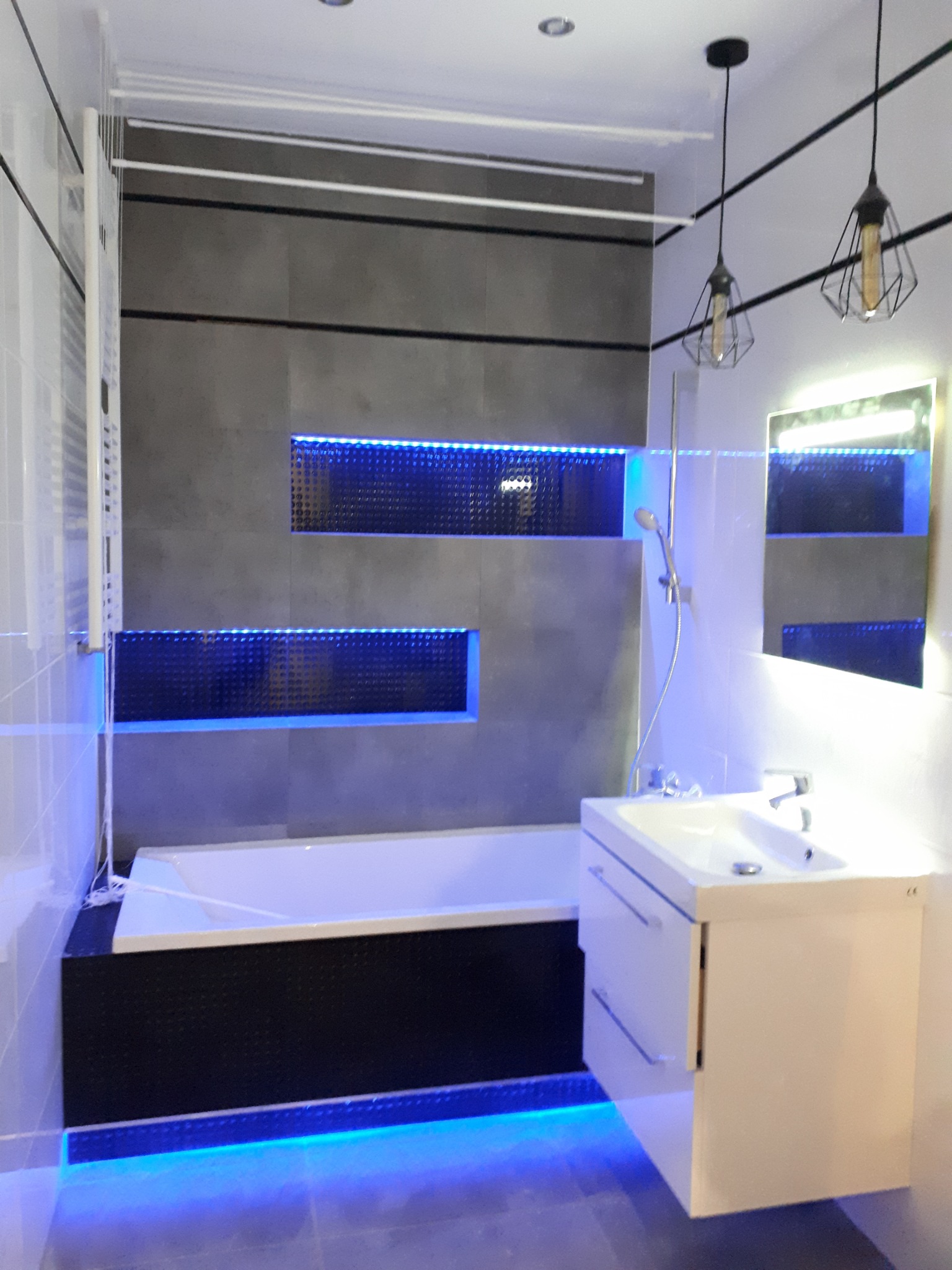 Nowoczesna łazienka z podświetlaną wanną, umywalką z szafką i geometrycznymi lampami wiszącymi na tle szarej ściany z wnękami oświetlonymi niebieskim światłem LED.