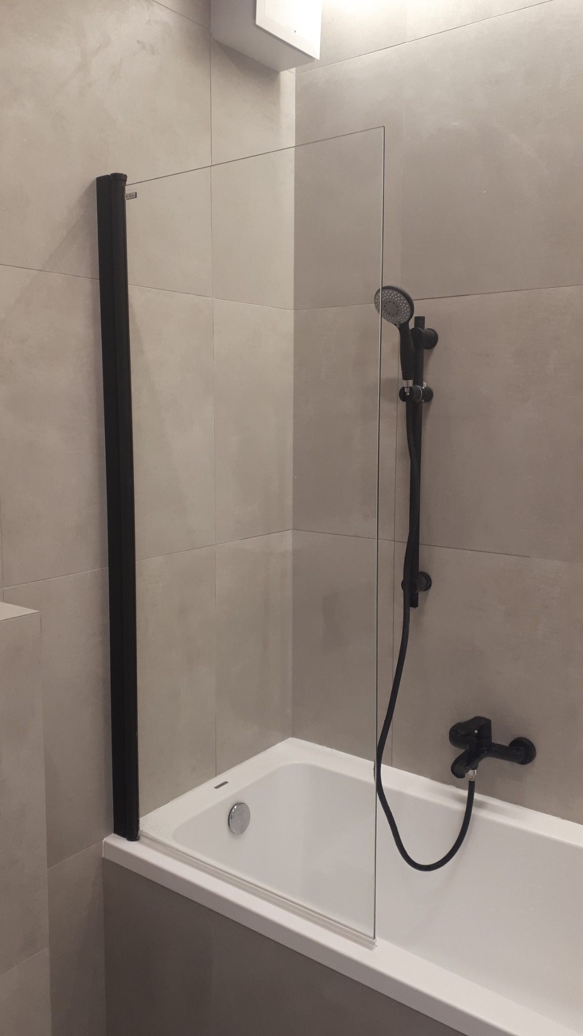 Nowoczesna łazienka z białą wanną, czarną armaturą i szklanym parawanem z czarną ramą, na tle płytek w odcieniach szarości.