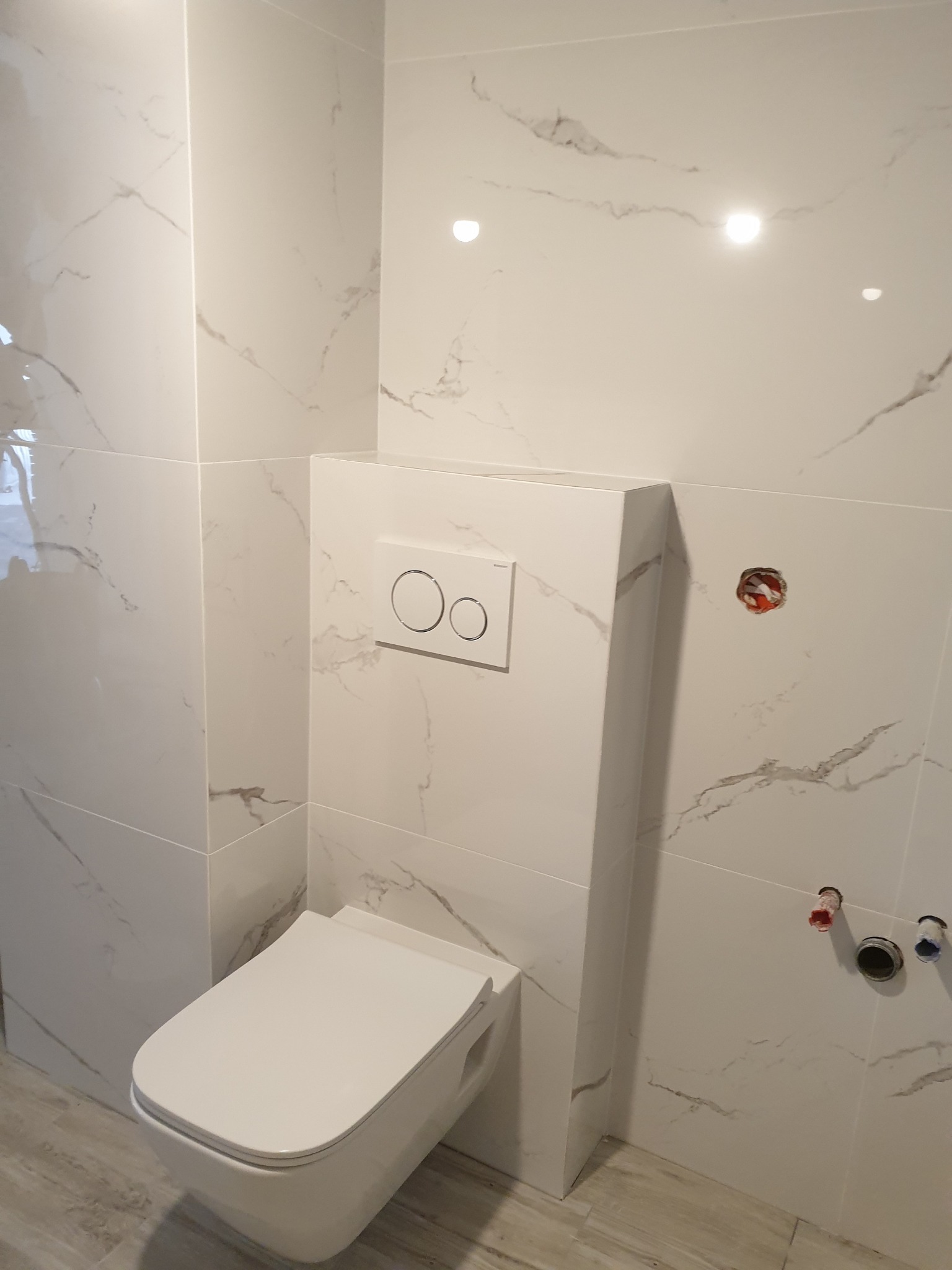 Nowoczesna, podwieszana toaleta z białymi płytkami imitującymi marmur, widoczne przygotowane instalacje wodne i elektryczne, szare płytki podłogowe.