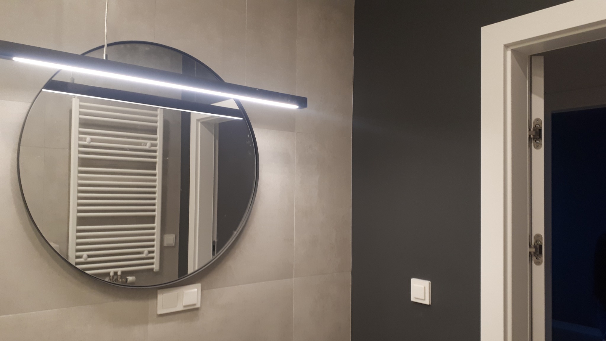 Nowoczesna łazienka z okrągłym lustrem z podświetleniem LED, widoczny grzejnik drabinkowy odbijający się w lustrze i szara ściana z białym włącznikiem światła.