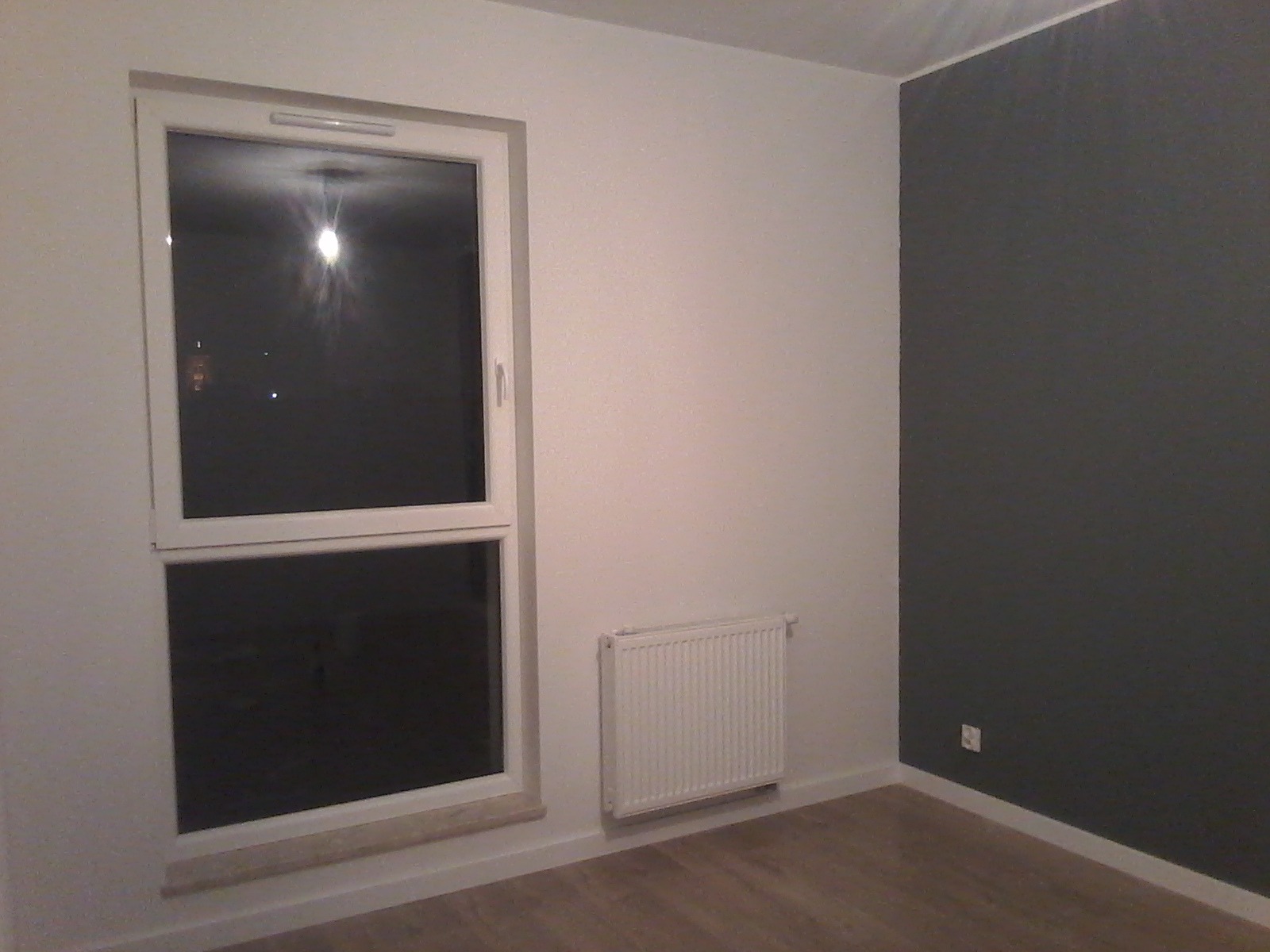 Pomalowane na dwa kolory wnętrze pokoju z białym oknem PCV, grzejnikiem i panelami podłogowymi w odcieniach drewna.