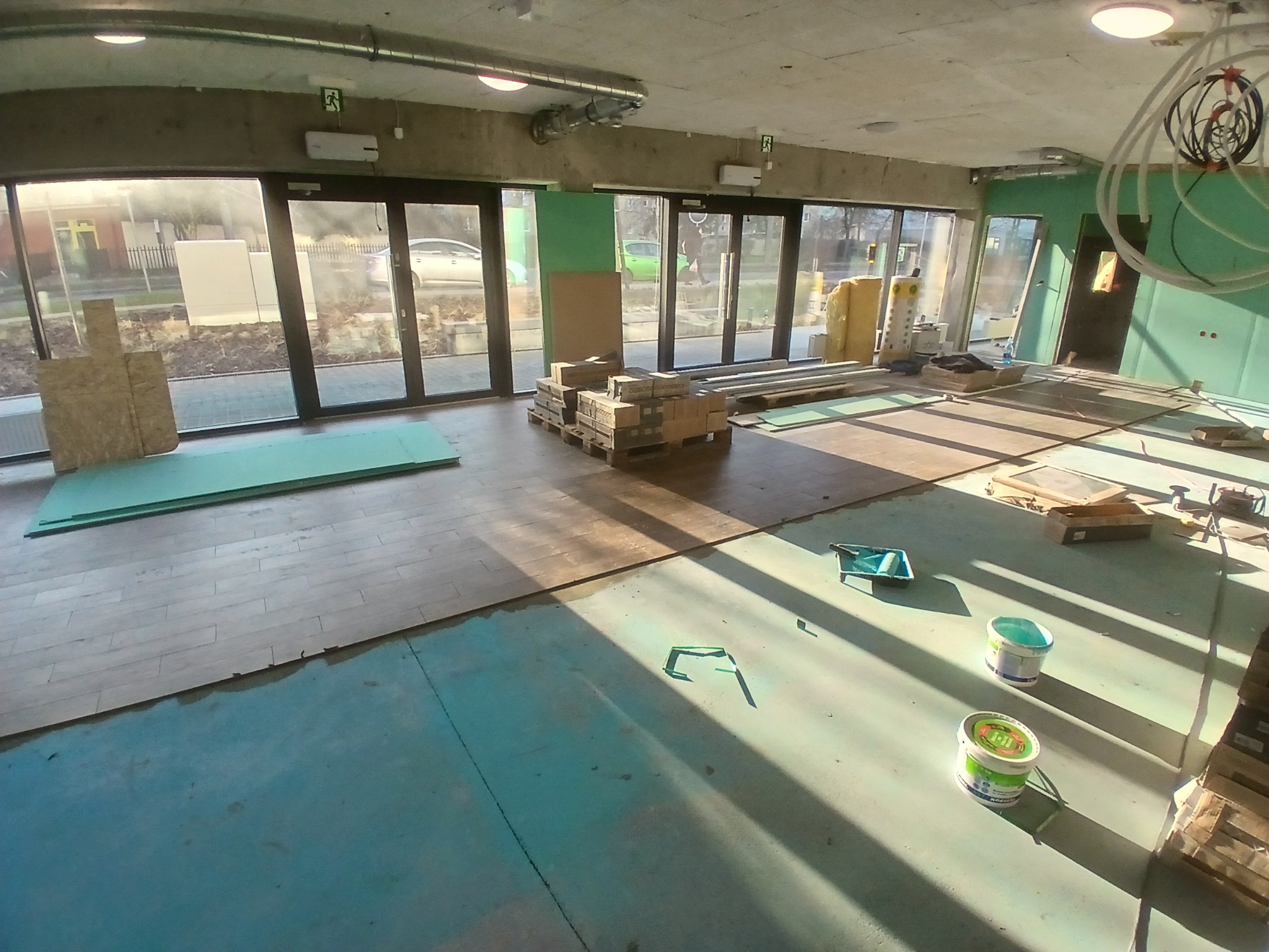 Wnętrze w trakcie prac, widoczne przygotowane panele podłogowe, puszki z farbą, płyty OSB i materiały izolacyjne, duże okna z widokiem na zewnątrz.