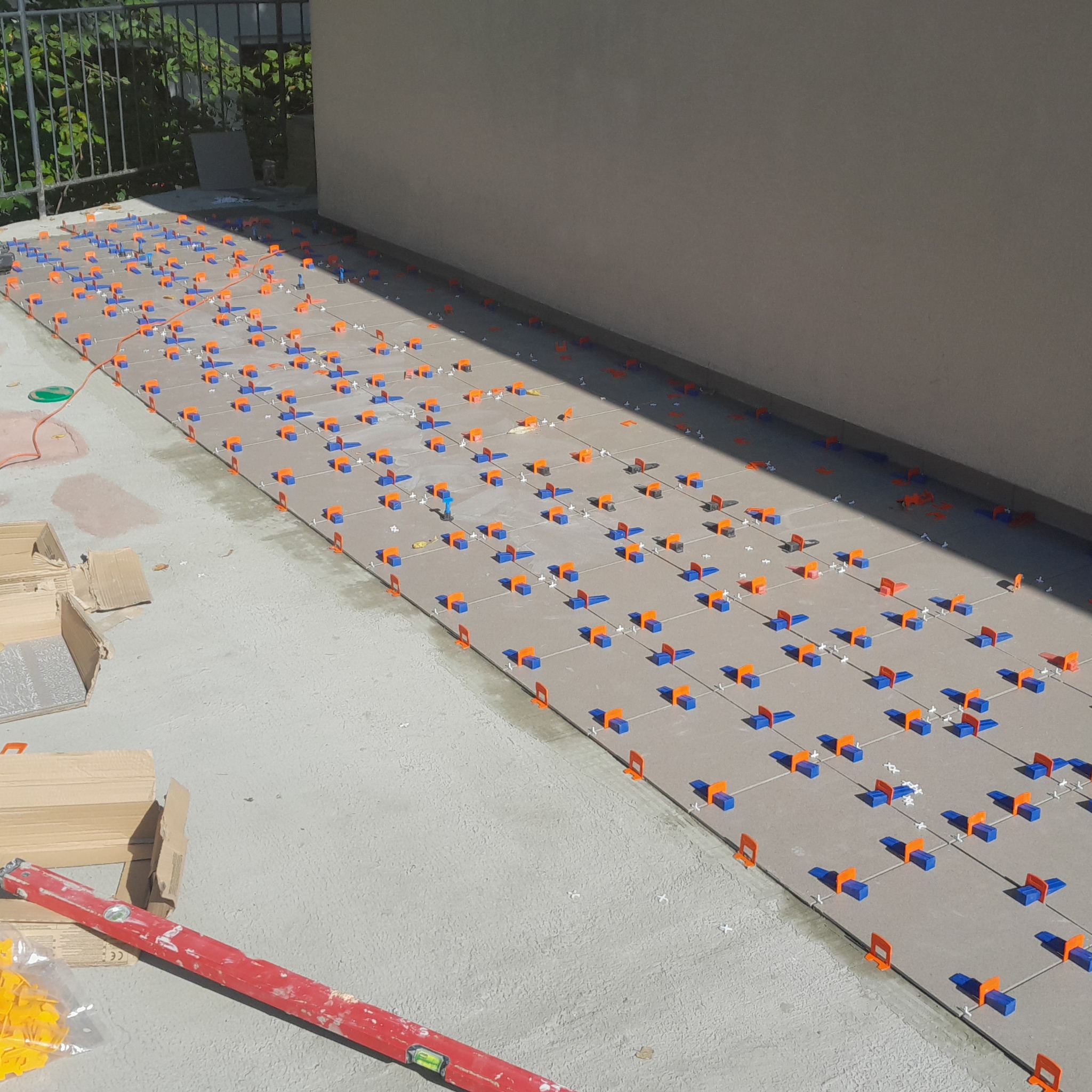 Układanie szarych płytek na tarasie z użyciem pomarańczowo-niebieskich klipsów poziomujących. Widoczna poziomica i kartony z płytkami.