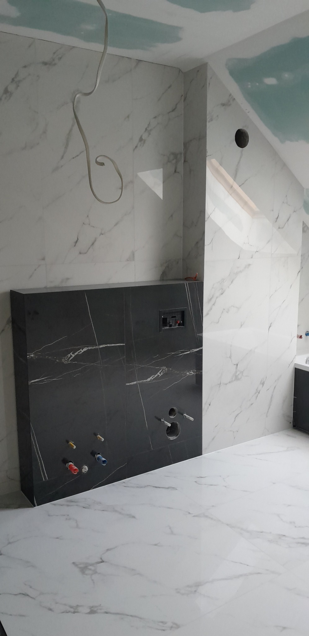 Wykończona ściana w łazience na poddaszu z białymi i czarnymi płytkami imitującymi marmur. Widoczne instalacje wodne i elektryczne, oraz fragment sufitu z płyt g-k.