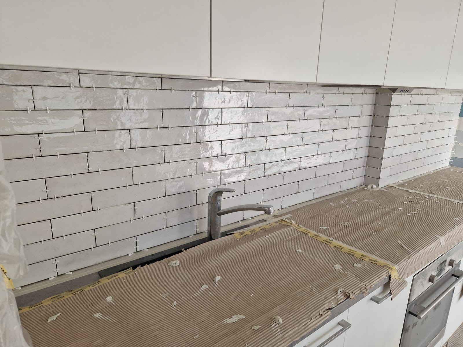 Układanie błyszczących, beżowych płytek w kuchni. Widoczne krzyżyki dystansowe. Zabezpieczony blat i białe szafki kuchenne. Kran zamontowany.