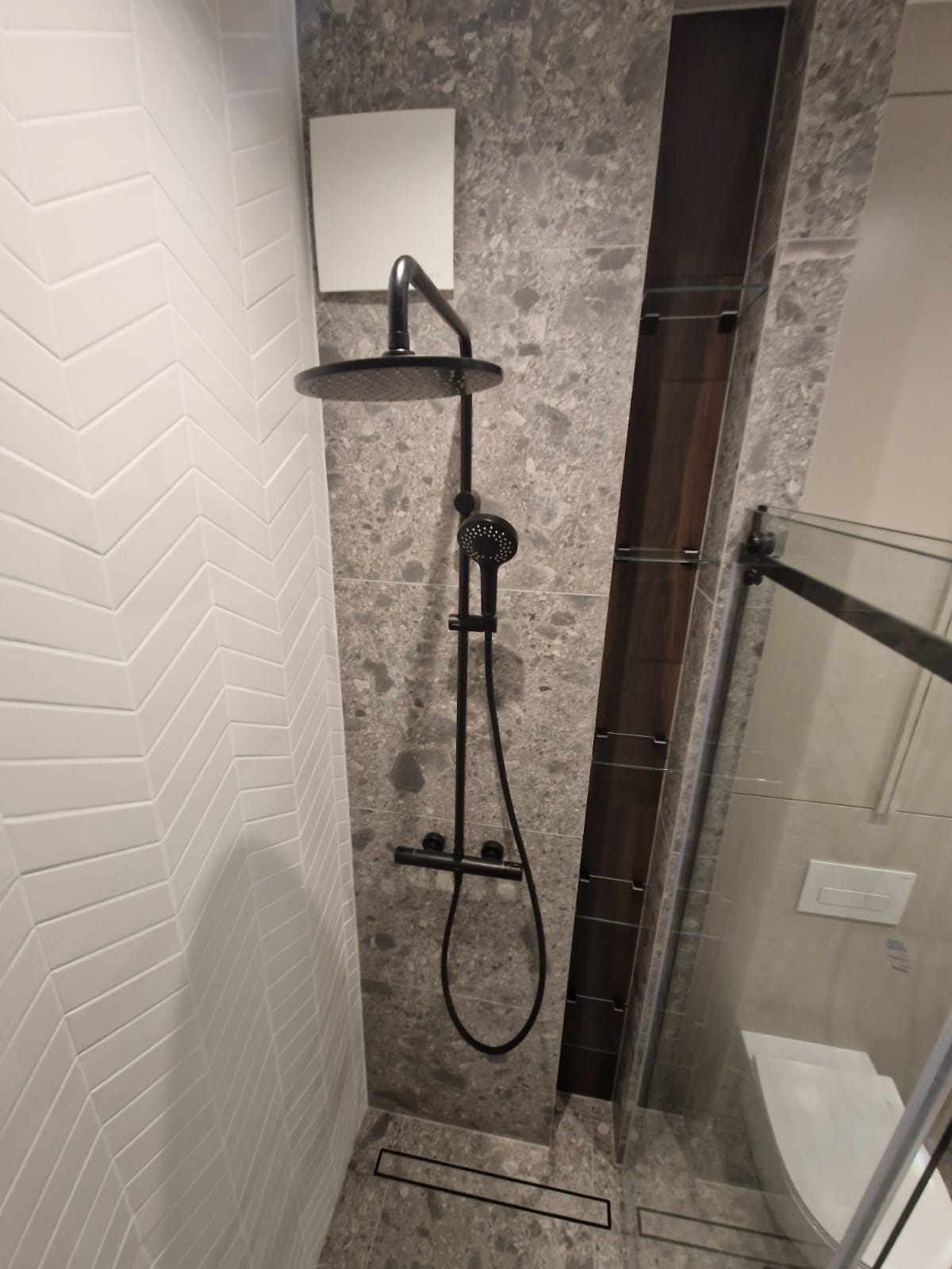 Nowoczesna kabina prysznicowa z deszczownicą i białymi płytkami ułożonymi w jodełkę, szare płytki imitujące kamień, czarna armatura. Minimalistyczny design.