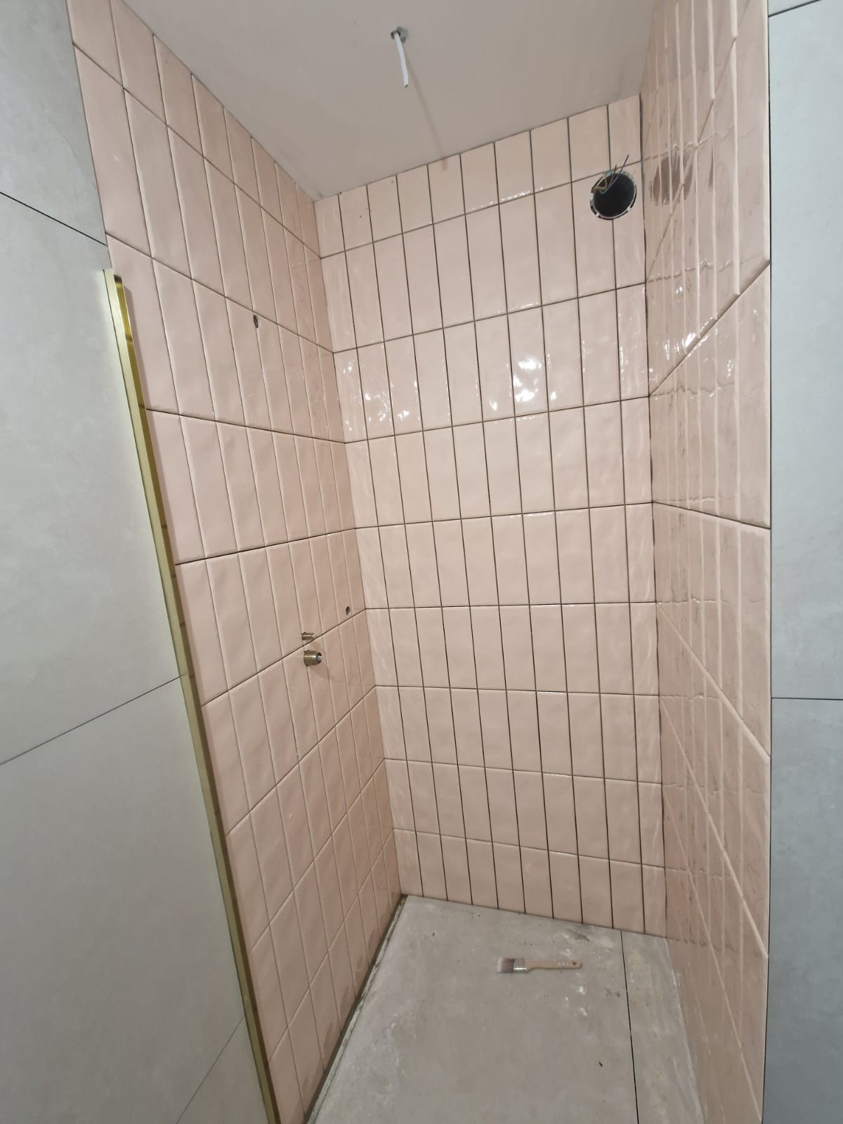 Ściany kabiny prysznicowej wyłożone błyszczącymi, pionowymi, różowymi płytkami. Widoczne otwory na armaturę i niedokończona podłoga. W rogu leży pędzel.