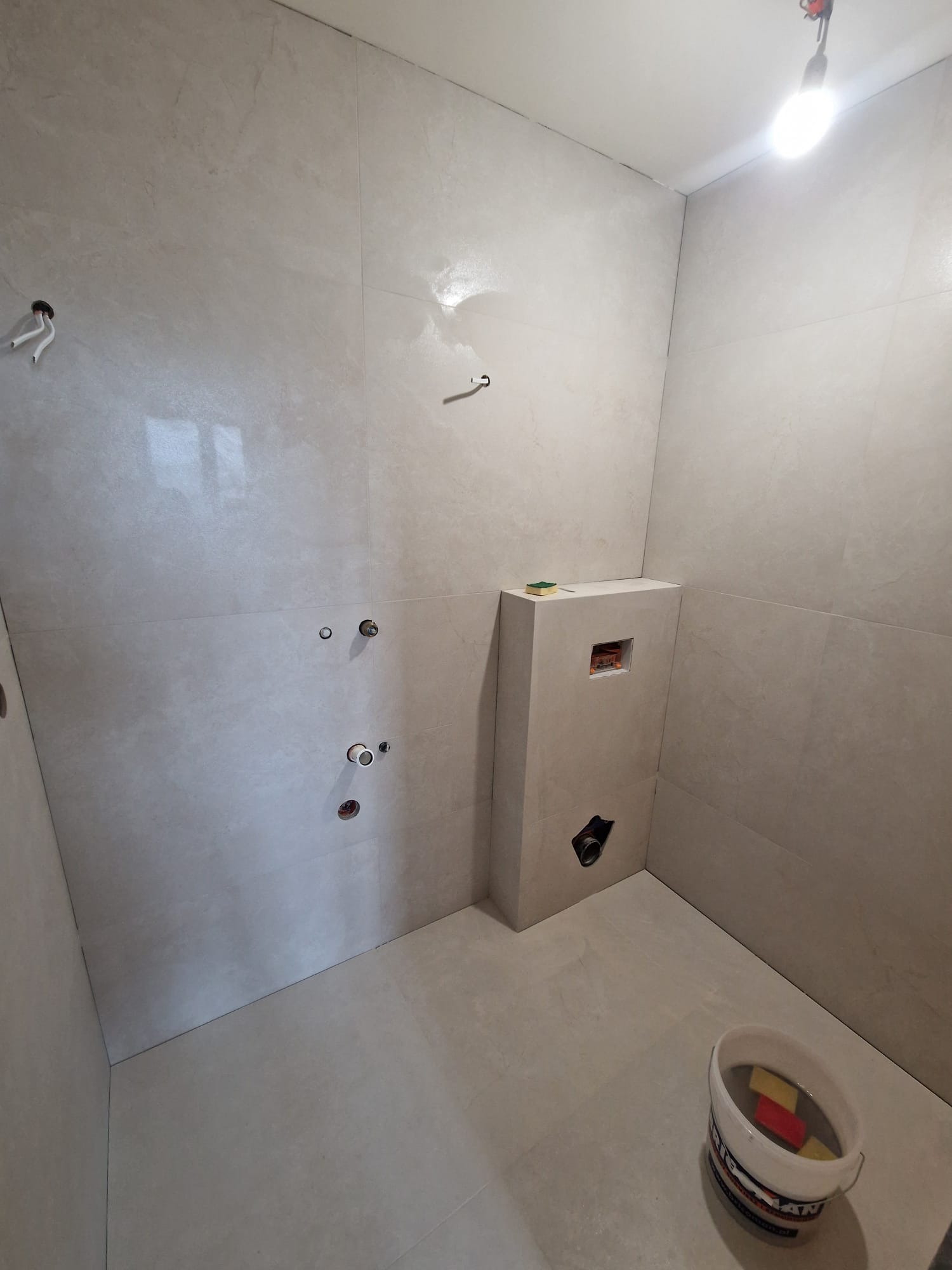 Jasna łazienka po remoncie z beżowymi płytkami, widoczne przyłącza wodne, fragment toalety podtynkowej i wiadro z gąbkami. Ukończone prace wykończeniowe.