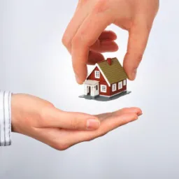 Dwie dłonie prezentują model domu z czerwonymi ścianami i oliwkowym dachem na tle jasnoszarego tła.