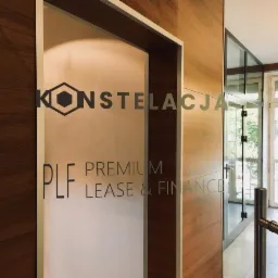 Szklane drzwi wejściowe do biura z napisem 'KONSTELACJA PLF PREMIUM LEASE & FINANCE' oraz logo w kształcie heksagonu z wpisanym sześcianem, widok z korytarza z drewnianą okładziną.
