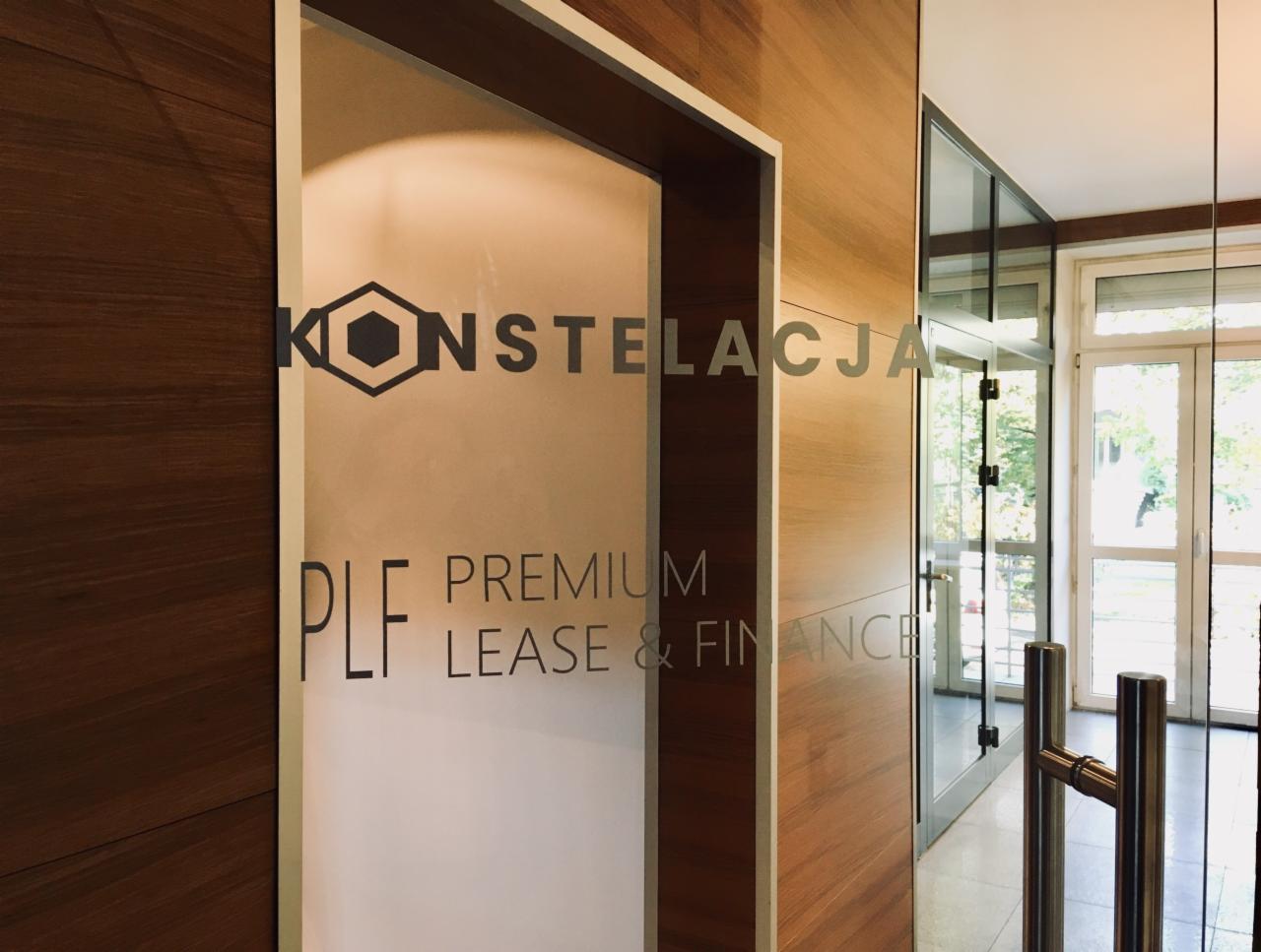 Szklane drzwi wejściowe do biura z napisem 'KONSTELACJA PLF PREMIUM LEASE & FINANCE' oraz logo w kształcie heksagonu z wpisanym sześcianem, widok z korytarza z drewnianą okładziną.