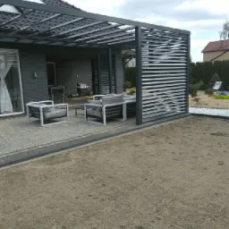 Nowoczesna, szara pergola z poziomymi lamelami, częściowo osłaniająca taras z meblami ogrodowymi; widoczne szare kostki brukowe i ziemia, w tle dom z szarą elewacją.