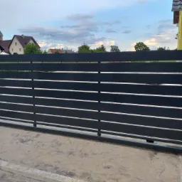 Nowoczesna, przesuwna brama wjazdowa z poziomych, grafitowych profili, widok z zewnątrz posesji, z domami w tle.