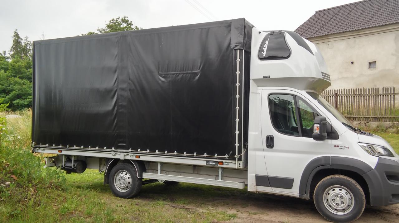 Biały bus z czarną plandeką na trawiastym terenie, widok z boku, na zabudowaniach widoczne logo 'LAMAR'.