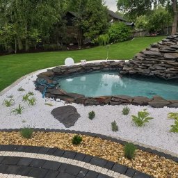 Zielone ogrody - Oczko wodne z kaskadą z kamienia łupkowego, otoczone białym i brązowym żwirem, z nasadzeniami roślin. W tle trawnik i drzewa.