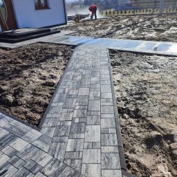 Zielone ogrody - Układanie kostki brukowej na posesji domu jednorodzinnego. Widoczny fragment ścieżki z kostki, ziemia i pracownik z taczką w tle. Słoneczny dzień.