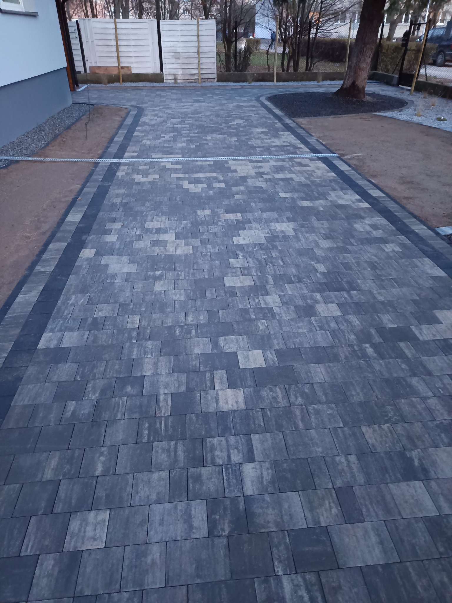 Ułożona kostka brukowa w odcieniach szarości tworząca ścieżkę i podjazd. Widoczne obrzeża i elementy dekoracyjne z kamieni. Tło stanowią budynek i drzewa.