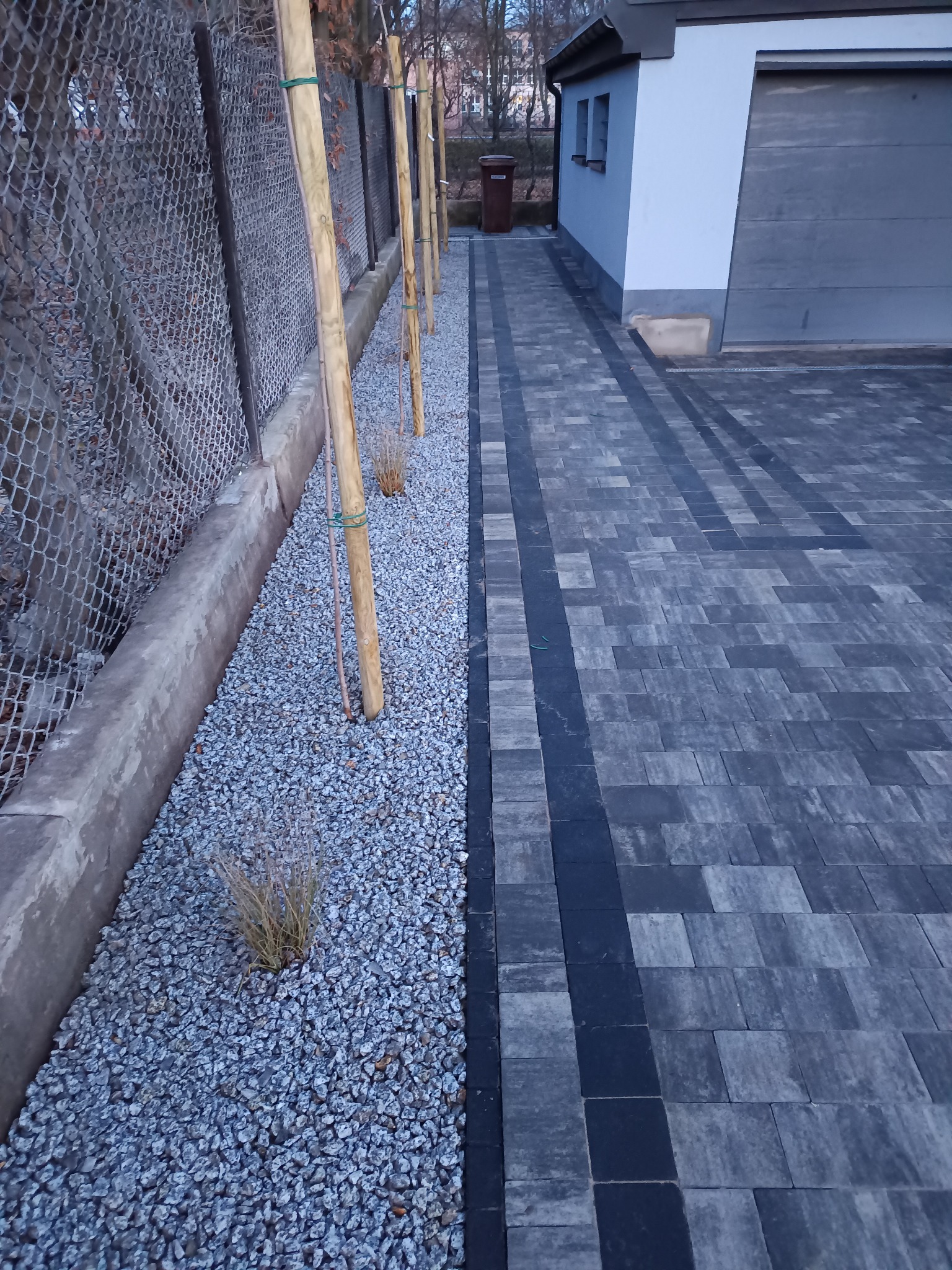 Świeżo posadzone drzewka wzdłuż ogrodzenia, zabezpieczone palikami, z drobnym żwirem i kostką brukową. Widok na budynek z garażem w tle.