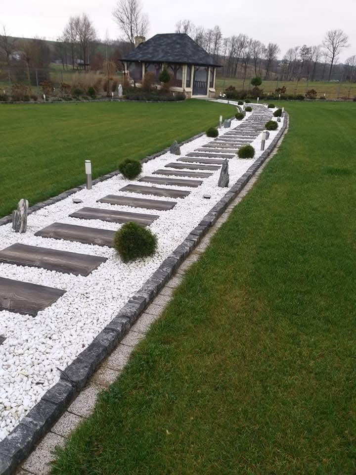 Ogród z ścieżką z drewnianych płyt i białego żwiru, prowadzącą do altany. Starannie przystrzyżony trawnik i dekoracyjne krzewy. Kamienne obrzeża ścieżki.