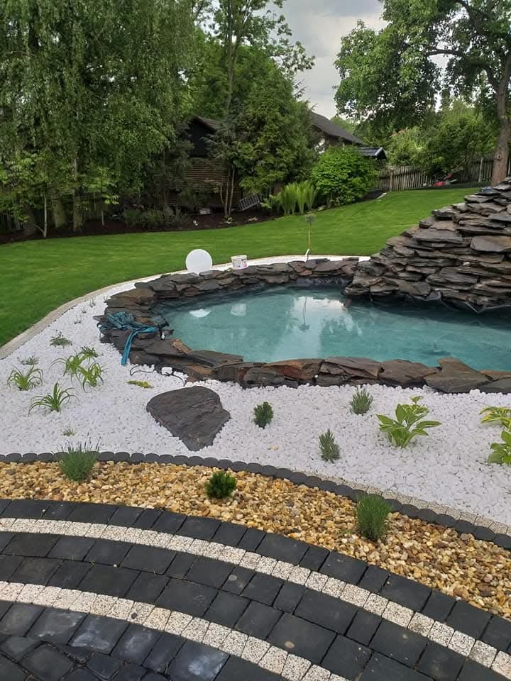 Oczko wodne z kaskadą z kamienia łupkowego, otoczone białym i brązowym żwirem, z nasadzeniami roślin. W tle trawnik i drzewa.