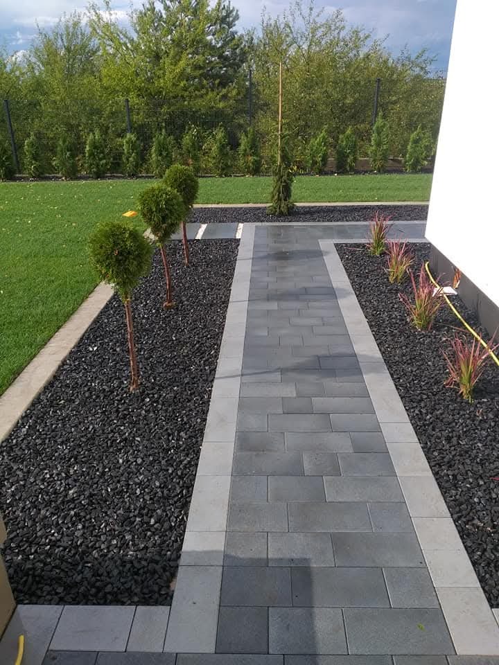 Ogród z ścieżką z szarych płyt, obłożoną czarnym grysem i ozdobnymi drzewkami. Estetyczne zagospodarowanie przestrzeni wokół domu.