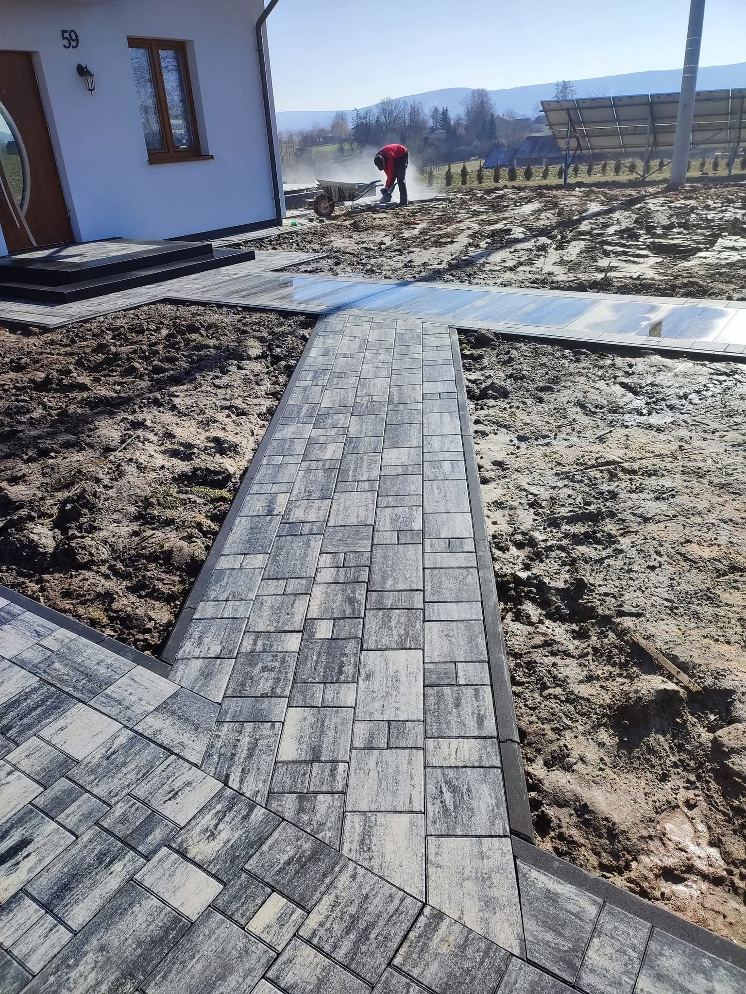 Układanie kostki brukowej na posesji domu jednorodzinnego. Widoczny fragment ścieżki z kostki, ziemia i pracownik z taczką w tle. Słoneczny dzień.