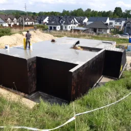 Wylany fundament pod budynek jednorodzinny z zabezpieczeniem przeciwwilgociowym, widoczny pracownik z łatą, w tle zabudowa mieszkalna.
