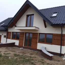 Nowoczesny, dwukondygnacyjny dom z czarną dachówką, drewnianymi elementami elewacji i częściowo zagospodarowanym terenem wokół.