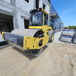 Żółty walec drogowy marki Bomag ubija żwirowe podłoże na placu budowy, w tle budynek w stanie surowym i palety kostki brukowej.