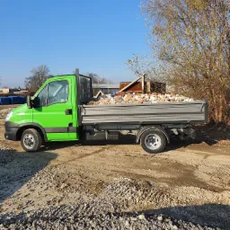 Jasnozielony samochód dostawczy z otwartą skrzynią ładunkową wypełnioną gruzem ceglanym, stojący na żwirowym podłożu w słoneczny dzień.