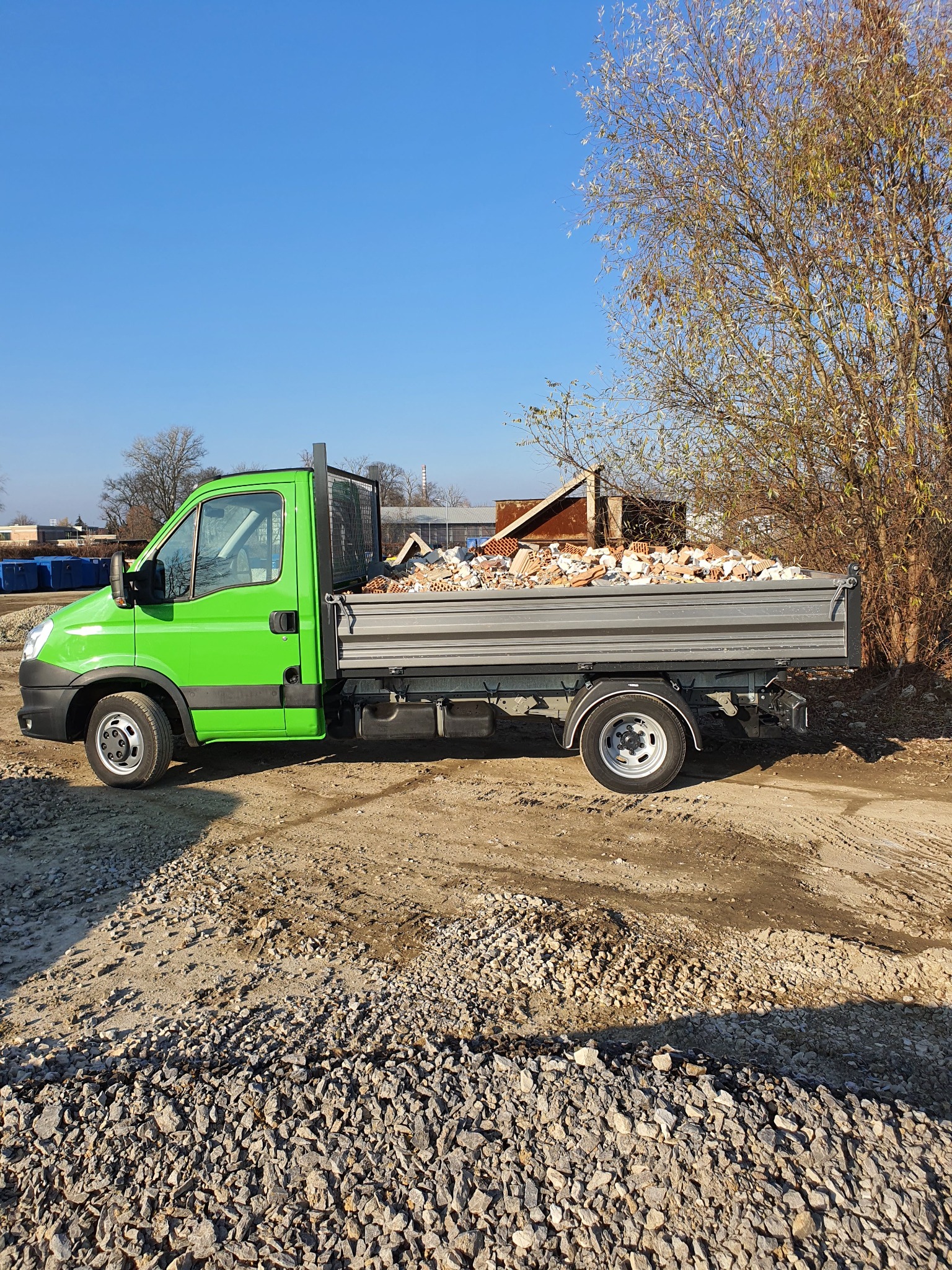 Jasnozielony samochód dostawczy z otwartą skrzynią ładunkową wypełnioną gruzem ceglanym, stojący na żwirowym podłożu w słoneczny dzień.