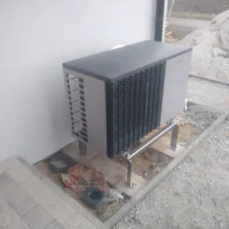 Zewnętrzna jednostka klimatyzacyjna z widocznymi przewodami elektrycznymi, zamontowana na metalowych wspornikach przy białej ścianie budynku, z hałdą piasku w tle.