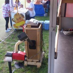 Ekowarm Roksana Panek - Mobilna instalacja grzewcza z czerwonym palnikiem, rurą i drewnianą obudową na trawie, obok stoiska z kukurydzą i ludźmi.