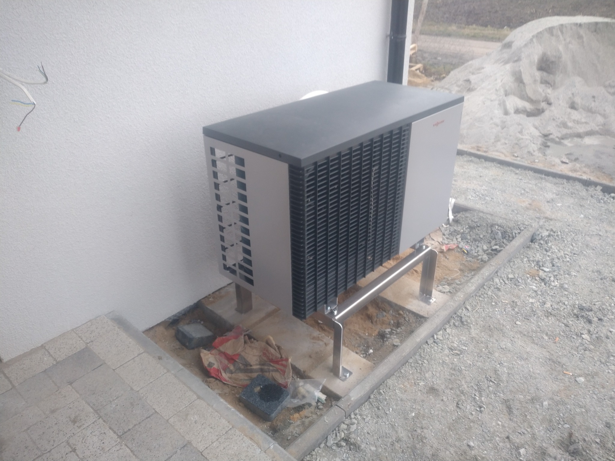 Zewnętrzna jednostka klimatyzacyjna z widocznymi przewodami elektrycznymi, zamontowana na metalowych wspornikach przy białej ścianie budynku, z hałdą piasku w tle.