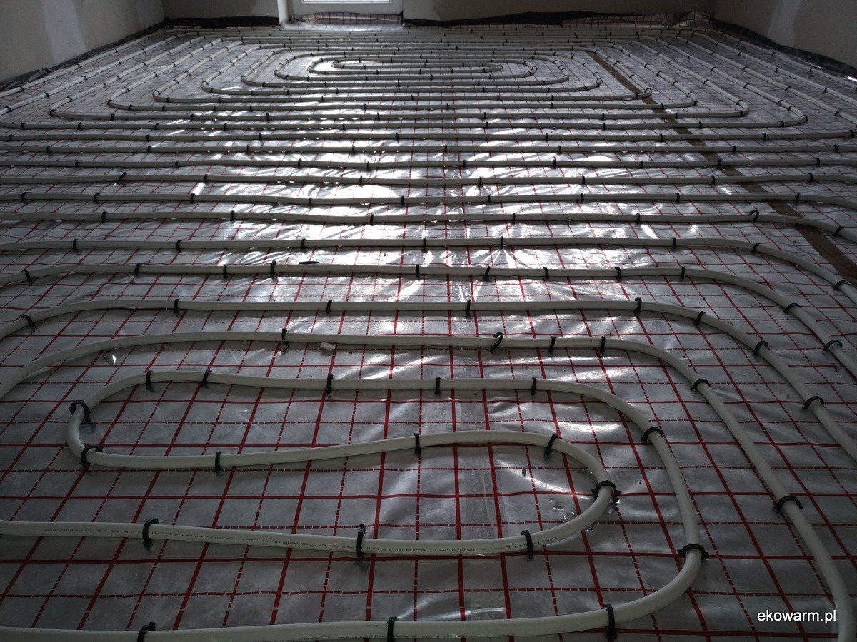 Rozłożone rury ogrzewania podłogowego na folii z siatką w nowym budynku, widoczne czarne uchwyty mocujące.