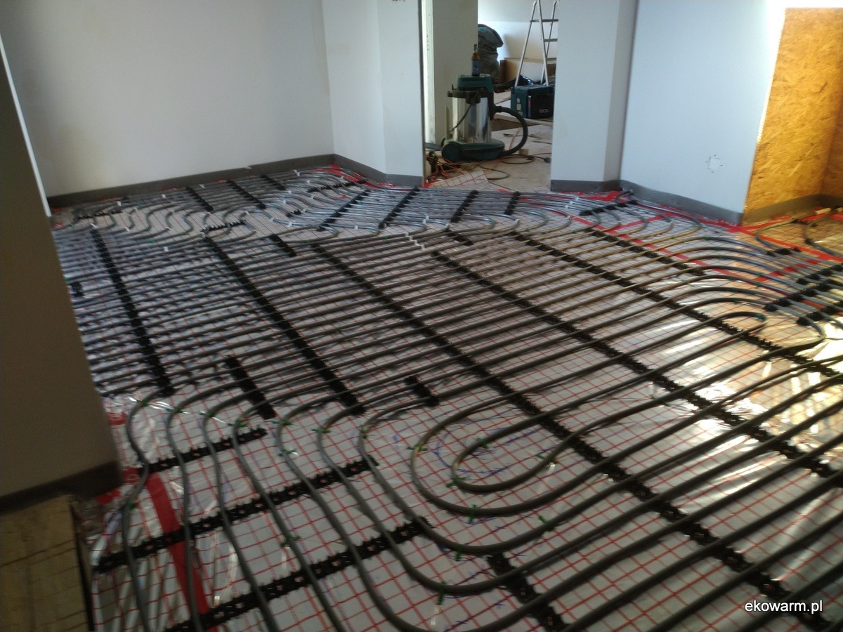 Montaż ogrzewania podłogowego: rury grzewcze ułożone na folii z siatką, widoczne narzędzia w tle.