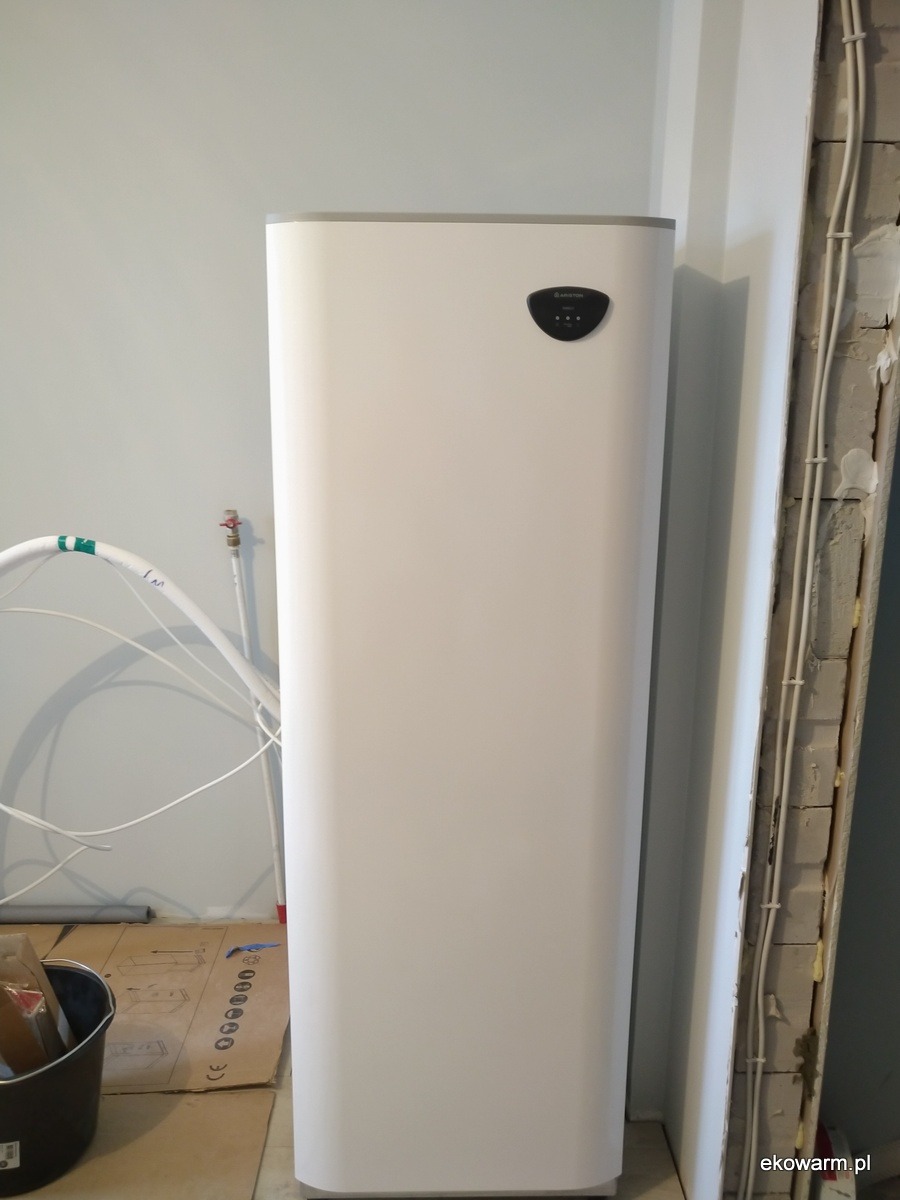 Biały, pionowy zbiornik z czarnym panelem sterowania, zamontowany przy ścianie w trakcie remontu, widoczne przewody i fragmenty niezabezpieczonej ściany.
