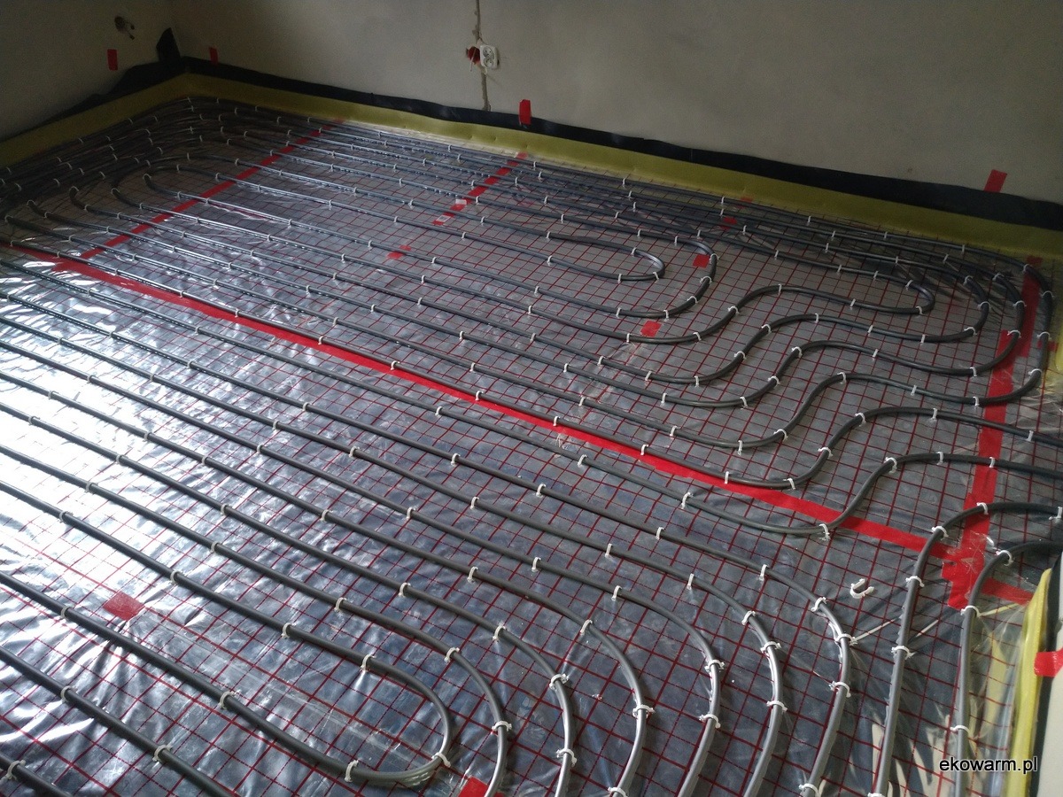 Instalacja ogrzewania podłogowego: rury grzewcze ułożone na folii z czerwoną siatką, przymocowane białymi uchwytami, widoczne fragmenty ścian w surowym stanie.