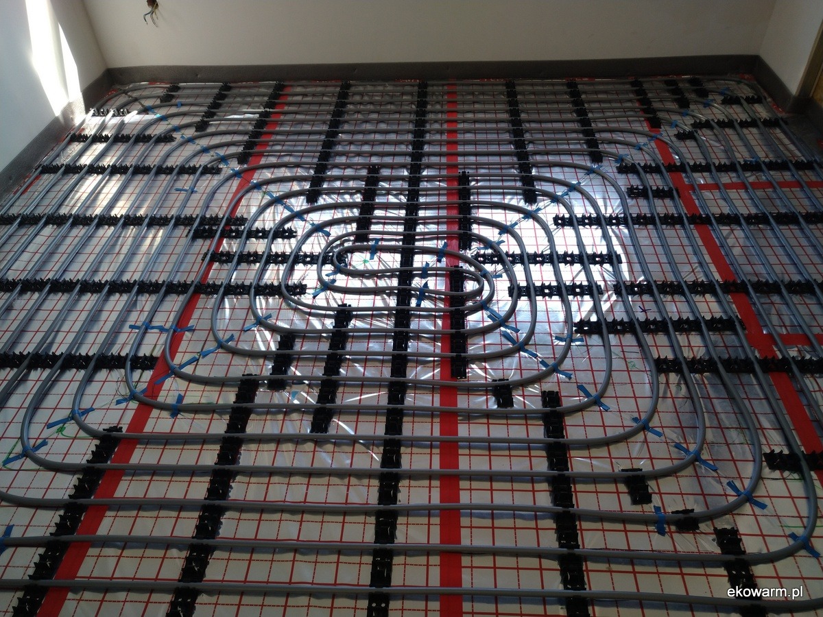 Układ rur ogrzewania podłogowego w trakcie montażu, widoczne czarne uchwyty mocujące i folia z czerwoną siatką, rury połączone niebieskimi taśmami.