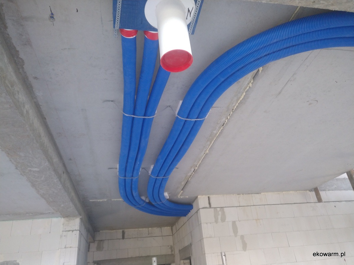 Niebieskie, karbowane rury wentylacyjne, częściowo przymocowane łańcuszkami do sufitu z betonu, widok instalacji rekuperacji w surowym wnętrzu.