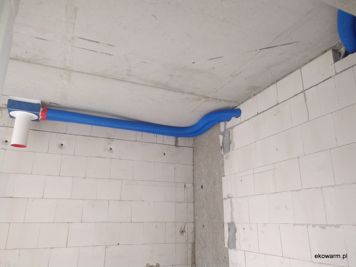 Niebieskie rury wentylacyjne w karbowanej osłonie, poprowadzone pod betonowym stropem w surowym wnętrzu budynku z białych bloczków.