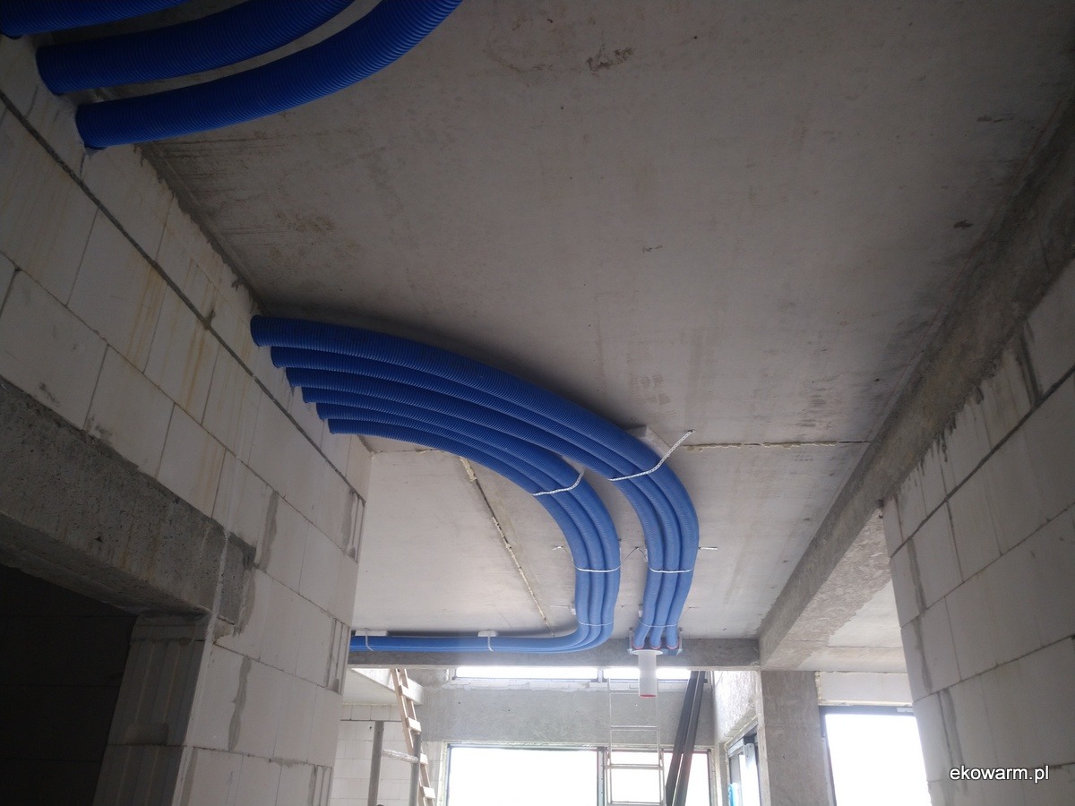 Instalacja wentylacji mechanicznej z rekuperacją w surowym wnętrzu budynku, widoczne niebieskie rury wentylacyjne przymocowane do sufitu i ścian z betonu komórkowego.
